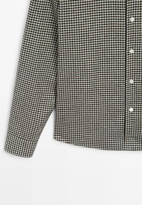 Camicia a quadri bianca e nera con maniche lunghe, davanti con bottoni e orlo arrotondato. Il tessuto ha una texture liscia con dettagli del motivo compatto.