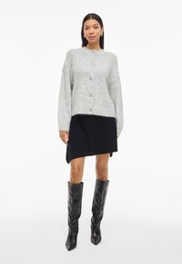 Šviesiai pilkas megztas kardiganas su apvaliu kaklu, sagčių uždarymu ir minkšta tekstūra; derinamas su juoda asimetriška suknele ir juodais ilgaaulių batais.