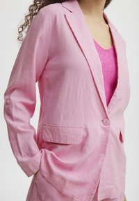 Pink linen blazer with a single button closure, notched lapels, and a front pocket. Worn over a pink knit top.