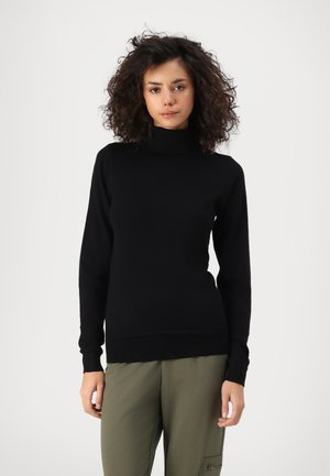 Jeune femme aux cheveux bouclés portant un pull à col roulé noir et un pantalon vert olive, debout devant un fond blanc uni.