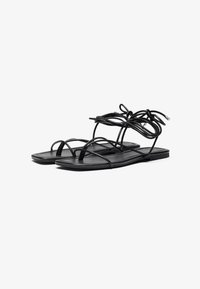 Sorte flate sandaler laget av glatt materiale, med flere tynne stropper og en knyttet detalj ved ankelen, åpen tå-design, minimalistisk stil.
