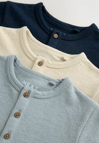 Trois chemises Henley en marine, crème et bleu clair. Chacune présente un col rond, une patte de boutonnage et un tissu texturé.
