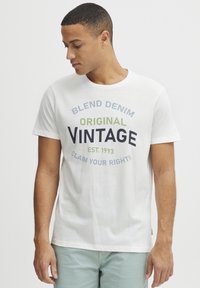 Hvit bomulls T-skjorte med rund hals, som har et fargerikt tekstdesign. Teksten inkluderer "BLEND DENIM," "ORIGINAL VINTAGE," og "CLAIM YOUR RIGHTS."