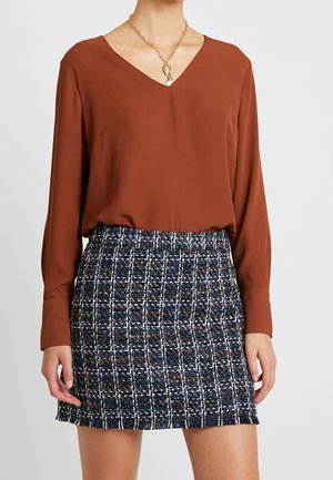 Vrouw draagt een roestbruine lange mouwen V-hals blouse, ingestopt in een marineblauwe en witte geruite tweed mini-rok met een gouden ketting.