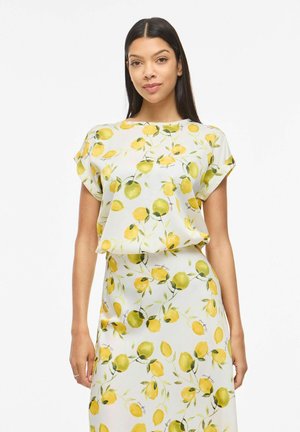 Kobieta w białej sukience z podwiniętymi rękawami i wzorem cytryn, stojąca na jednolitym białym tle.