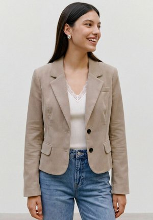 Jeune femme souriante portant un blazer beige en velours côtelé, un haut blanc orné de dentelle et un jean bleu, debout devant un fond clair uni.