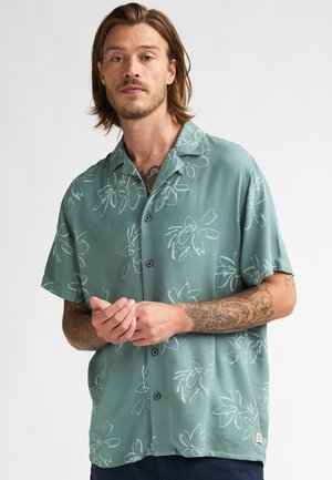 Camisa de manga corta de color verde claro con patrones florales blancos, cierre frontal con botones y botones negros. Textura suave y corte relajado.