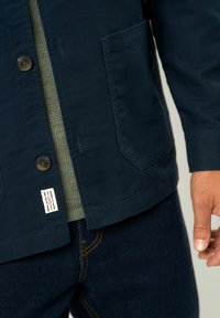 Chaqueta navy de algodón, con cierre de botones, bolsillo de parche y una etiqueta en el dobladillo. Lleva encima una camisa texturizada de color verde oliva.