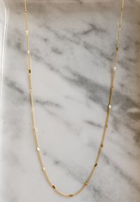 Collana in oro con maglie alternate piatte e testurizzate, con accenti a forma di fiore, esposta su uno sfondo di marmo.