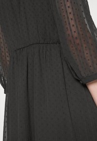 Svart klänning med genomskinliga puffärmar, rynkad midja och små, strukturerade prickar genom hela tyget som skapar ett subtilt mönster.