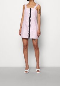 Robe à carreaux rose et blanc avec un décolleté carré, des bretelles à volants, une patte de boutonnage noire et des boutons décoratifs. Portée avec des sandales blanches à enfiler.