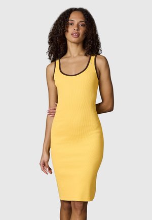 Frau mit lockigem Haar, die ein ärmelloses gelbes Kleid mit dunkelbraunem Besatz trägt, steht vor einem schlichten hellen Hintergrund.