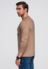 Langarmshirt in Beige mit rundem Ausschnitt und schwarzem grafischen Detail, kombiniert mit blauen Jeans, Model posiert seitlich und trägt eine schwarze Uhr.