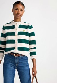 Zöld és krémszínű csíkos kardigán, kerek nyakkivágással, elöl gombokkal és egy arany szív részlettel. Farmerrel és barna övvel viselve.