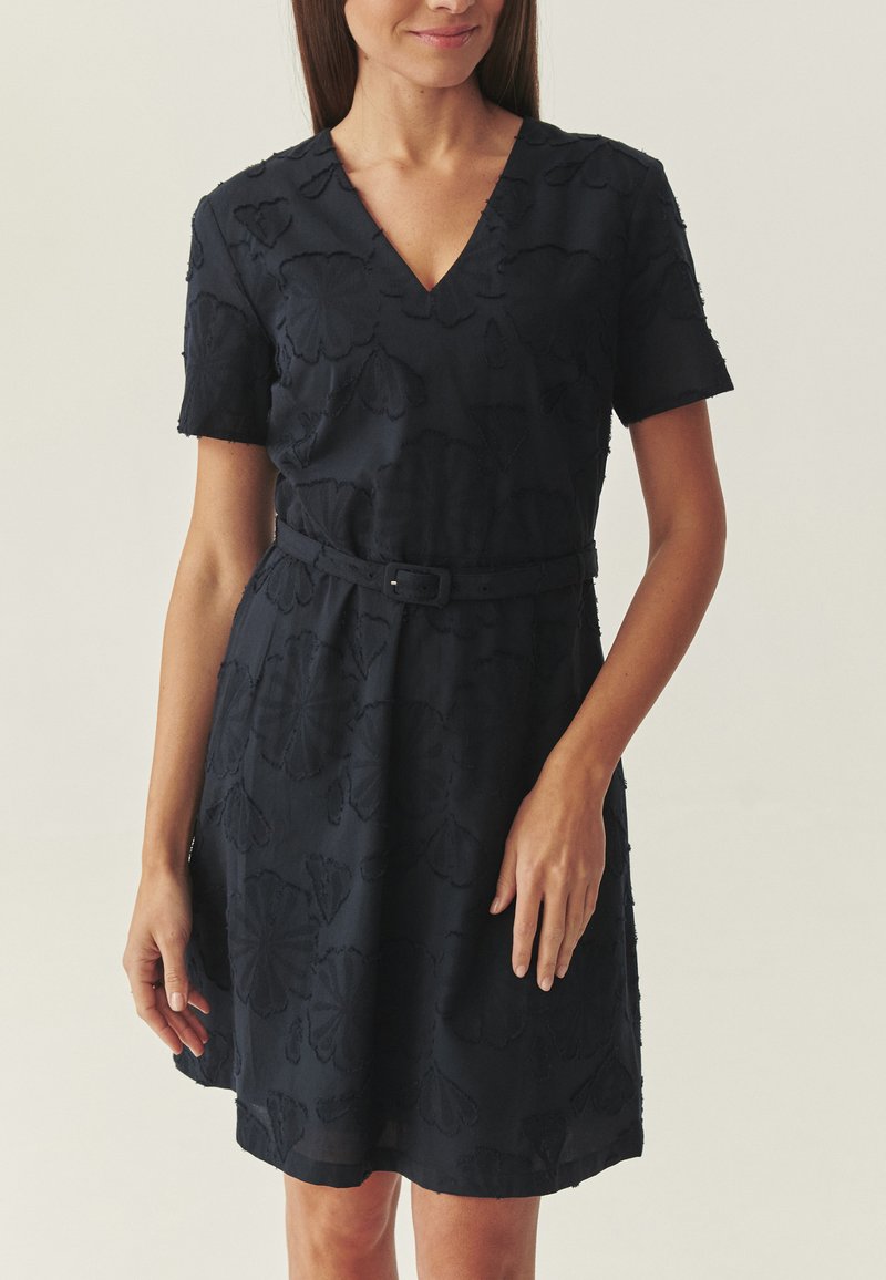 Donna che indossa un vestito nero al ginocchio con maniche corte, scollo a V, motivo floreale in rilievo e cintura abbinata in vita.