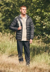 Doudoune noire avec texture matelassée sur un haut en polaire beige, associée à un pantalon noir. La personne tient une sangle de sac à dos et se tient sur l'herbe.