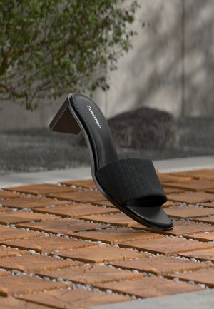 Black open-toe Calvin Klein mule with block heel on wooden tile floor, outdoor setting with blurred greenery and concrete background.