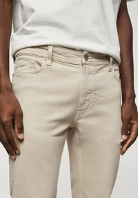Pantalones de algodón beig con un corte recto, diseño de cinco bolsillos y cierre de botón metálico. Textura suave y detalles de costura mínimos.