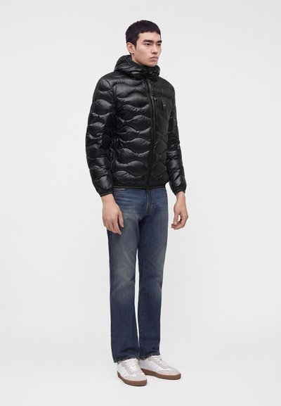 Jeune homme debout portant une veste matelassée à capuche noire, un jean bleu et des baskets blanches sur un fond clair uni.