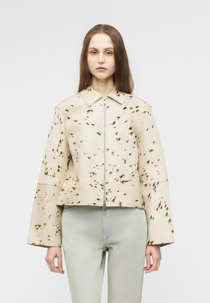 Γυναίκα με ίσια καστανά μαλλιά φοράει μπεζ και καφέ ρουστίκ μπουφάν με φερμουάρ και φαρδιά μανίκια, καθώς και ανοιχτό γκρι παντελόνι, στέκεται μπροστά σε απλό φόντο.