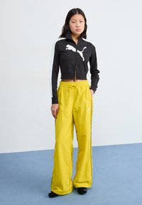Black cropped jacket with white accents and Puma logo, paired with loose yellow pants featuring black stripes and drawstring waist.