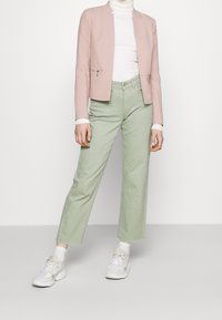 Světle růžový šitý blazer s detaily na zip, bílý žebrovaný rolák a zelené rovné kalhoty, doplněné bílými teniskami.