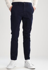 Chinos in cotone stretch blu navy con due tasche laterali e tasche posteriori, dotati di chiusura a bottone e design slim fit. Abbinati a sneakers bianche.