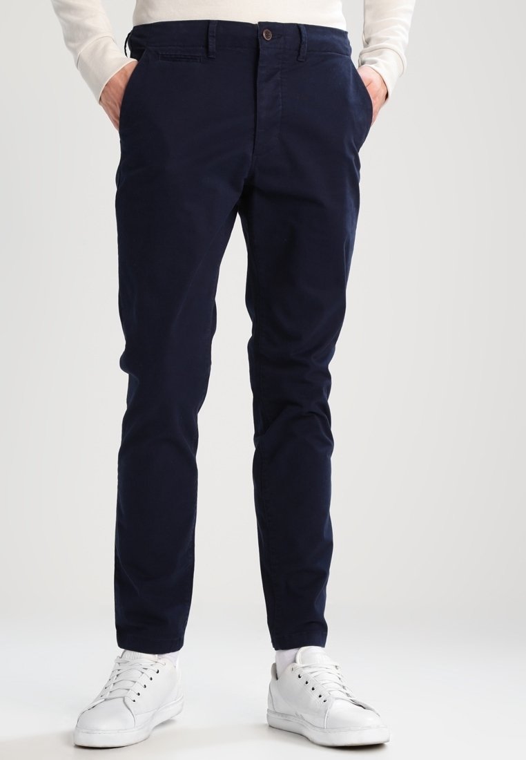 Chinos in cotone stretch blu navy con due tasche laterali e tasche posteriori, dotati di chiusura a bottone e design slim fit. Abbinati a sneakers bianche.