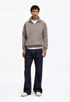 Jeune homme debout portant un sweat-shirt gris avec une fermeture éclair quart, un sous-t-shirt blanc, un jean sombre et ample, et des baskets blanches sur fond blanc.