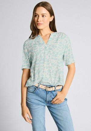 Femme aux longs cheveux bruns portant un chemisier à manches courtes bleu clair à motifs, rentré dans un jean bleu clair avec une ceinture beige.