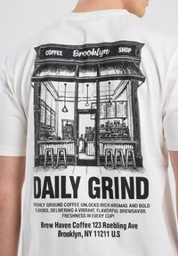 Camiseta blanca con un gráfico negro del interior de una cafetería con el texto "DAILY GRIND" y "Brew Haven Coffee, Brooklyn, NY."
