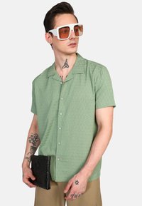 Camisa de manga corta verde con un patrón texturizado, botón frontal y tejido suave. Combinada con un clutch negro texturizado y gafas de sol blancas de gran tamaño.