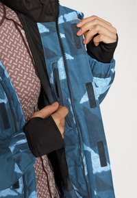 Giacca blu a fantasia con chiusure a zip e velcro, indossata sopra una camicia mauve texture. Presenta polsini neri e fodera interna.