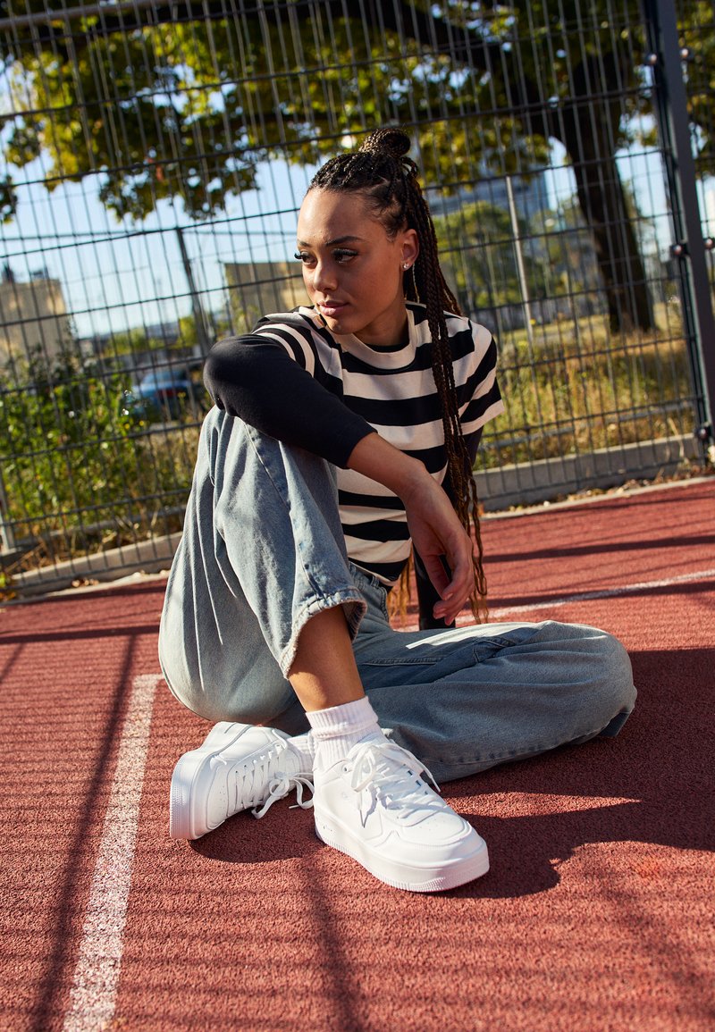 Sneakers bianchi con superficie testurizzata e suola spessa. Jeans in denim con orli arrotolati e camicia a maniche lunghe a righe bianche e nere. Ambientazione all'aperto.