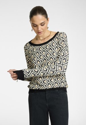 Maglione a maniche lunghe color crema con motivo geometrico a diamante nero e oro, rifinito con polsini e orlo a coste neri. Jeans neri.