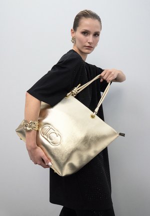 Bolso tote metálico dorado con acabado texturizado, doble asa superior y detalle del logo. La modelo lleva una blusa negra y un accesorio decorativo en la muñeca.