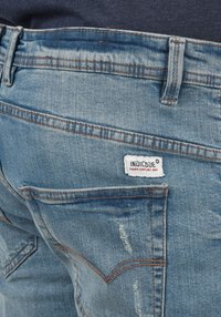 Lys blå denim jeans med en falmet finish, synlig sømdetalj i orange, og en hvit merkevareetikett som leser "INDICODE" på baklommen.