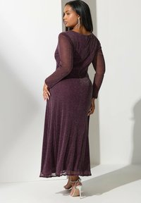 Lång lila klänning med långa genomskinliga ärmar, en figurnära bodice och en svävande kjol. Texturerad tyg med en subtil lyster. Silverfärgade högklackade skor.