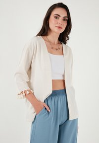 Giacca stile kimono beige chiaro con dettagli in frange sulle maniche, indossata sopra un top corto bianco e pantaloni larghi blu, che mostrano un fit rilassato.