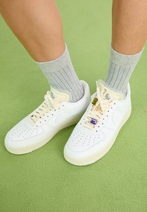 Adidași Nike Air Force 1 din piele albă, cu vârf perforat, șireturi crem și accente decorative de bijuterii. Sosete gri cu model în dungi vizibile. Fundal verde.