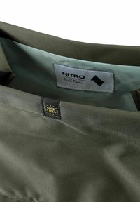 Interior de chaqueta verde oliva con forro azul claro, que presenta una etiqueta negra con "GOOD TIMES" y una etiqueta blanca marcada como "NITRO."