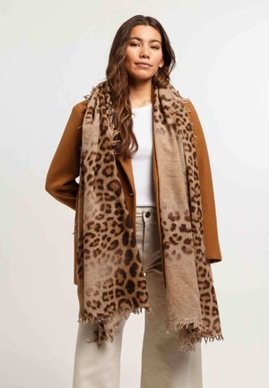 Sciarpa con stampa leopardata in beige e marrone. Tessuto morbido, bordi frangiati, appoggiata su un cappotto marrone, abbinata a una maglietta bianca e pantaloni chiari.