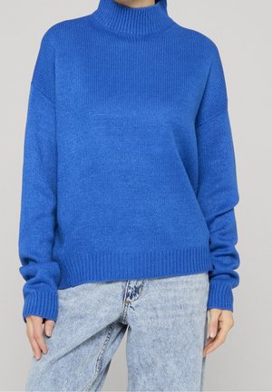 Personne portant un pull à col roulé côtelé bleu vif et un jean taille haute bleu clair, se tenant devant un fond uni.