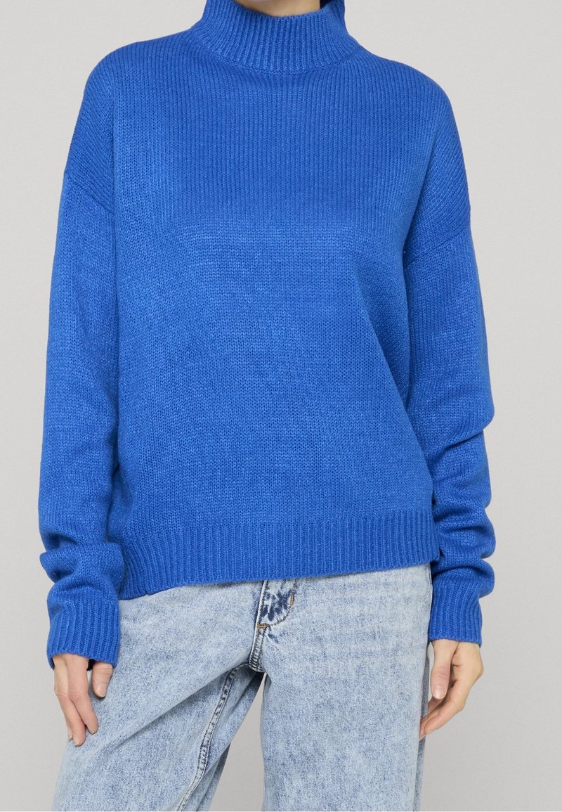 Personne portant un pull à col roulé côtelé bleu vif et un jean taille haute bleu clair, se tenant devant un fond uni.