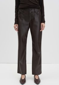 Woman wearing dark brown faux leather straight-leg pants and matching pointed-toe shoes, standing against a plain white background.