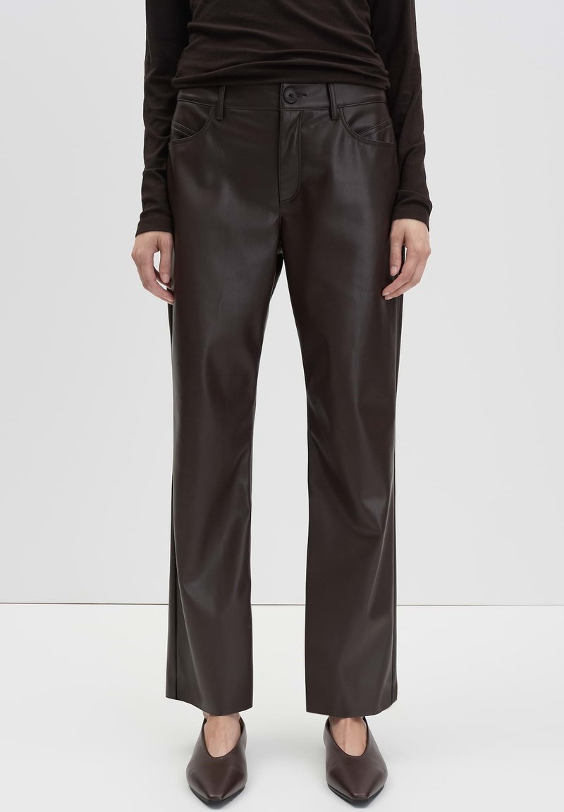 Woman wearing dark brown faux leather straight-leg pants and matching pointed-toe shoes, standing against a plain white background.