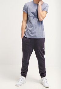 Šedé tričko s ohrnutými rukávy a námořnickým designem hvězdy, doplněné tmavě fialovými tepláky s dvěma kapsami a bílými teniskami.