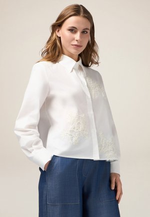 Camicia bianca con bottoni e maniche lunghe, caratterizzata da un intricato ricamo floreale bianco sulla parte anteriore. Abbinata a pantaloni blu a gamba larga.