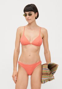 Koralni bikini s rebrastim uzorkom, podesivim trakama i donjim dijelom niskog struka. Model drži prugasti pleteni plažni ručnik. Prilozi uključuju ovalne sunčane naočale.