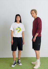 Sort cargo shorts og hvid grafisk t-shirt på den ene model; rød sweater på den anden. Begge bærer sneakers. Neutral baggrund og grøn gulv.